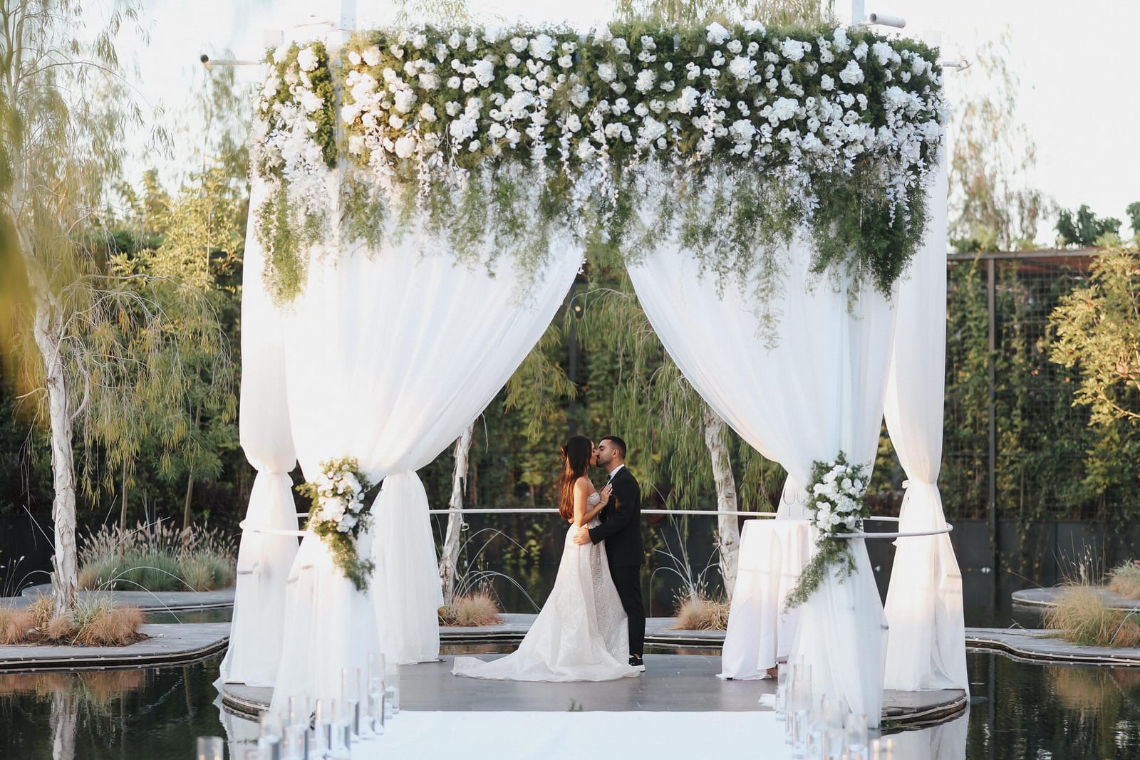 A couple embracing beneath a white chuppah topped with flowers on a platform in a pond, surrounded by willows
