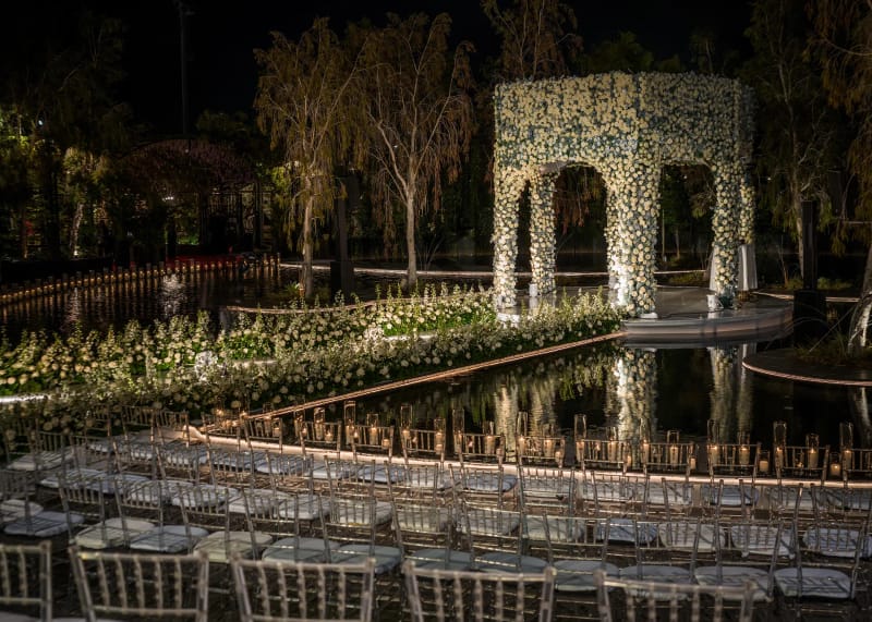 Pavillon de fleurs blanches au bord du bassin la nuit, avec rangées de chaises transparentes et bougies le long du bord