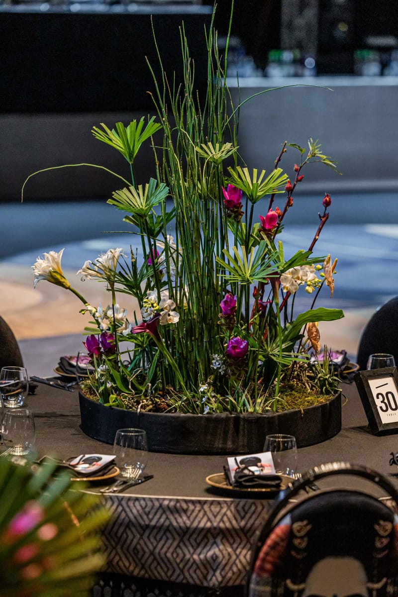 Plant arrangement in a round black planter with tall grasses, purple and white flowers and green leaves on a set table
