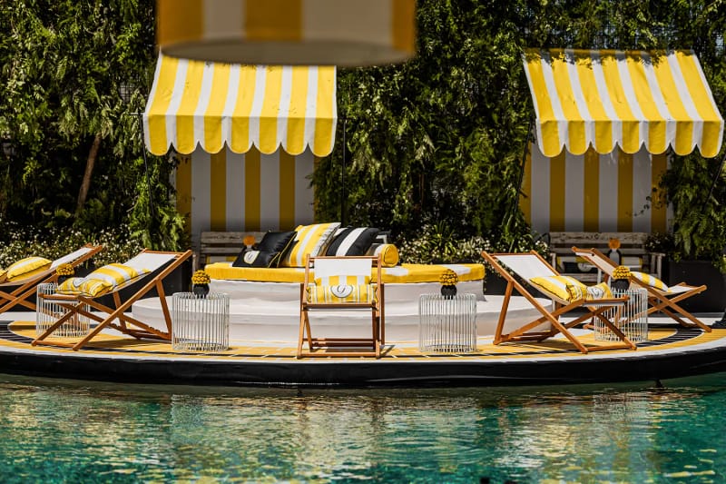 Two yellow and white striped cabanas with deck chairs and sofas on a poolside platform in front of a green wall