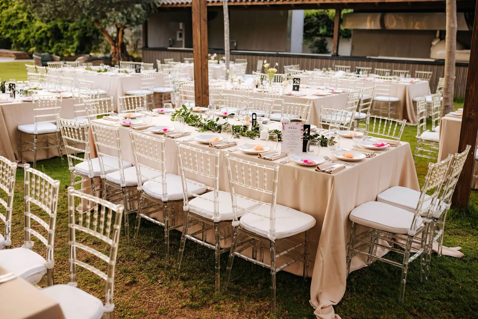 Longues tables aux nappes beiges et chaises transparentes à coussins blancs sur la pelouse, sous une pergola en bois