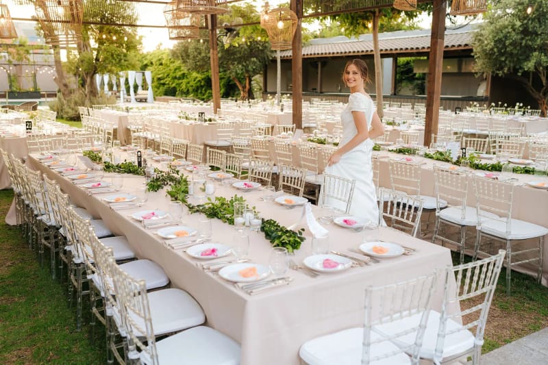 Une mariée debout près d'une longue table de banquet avec chemin de verdure, chaises transparentes et pergola en bois