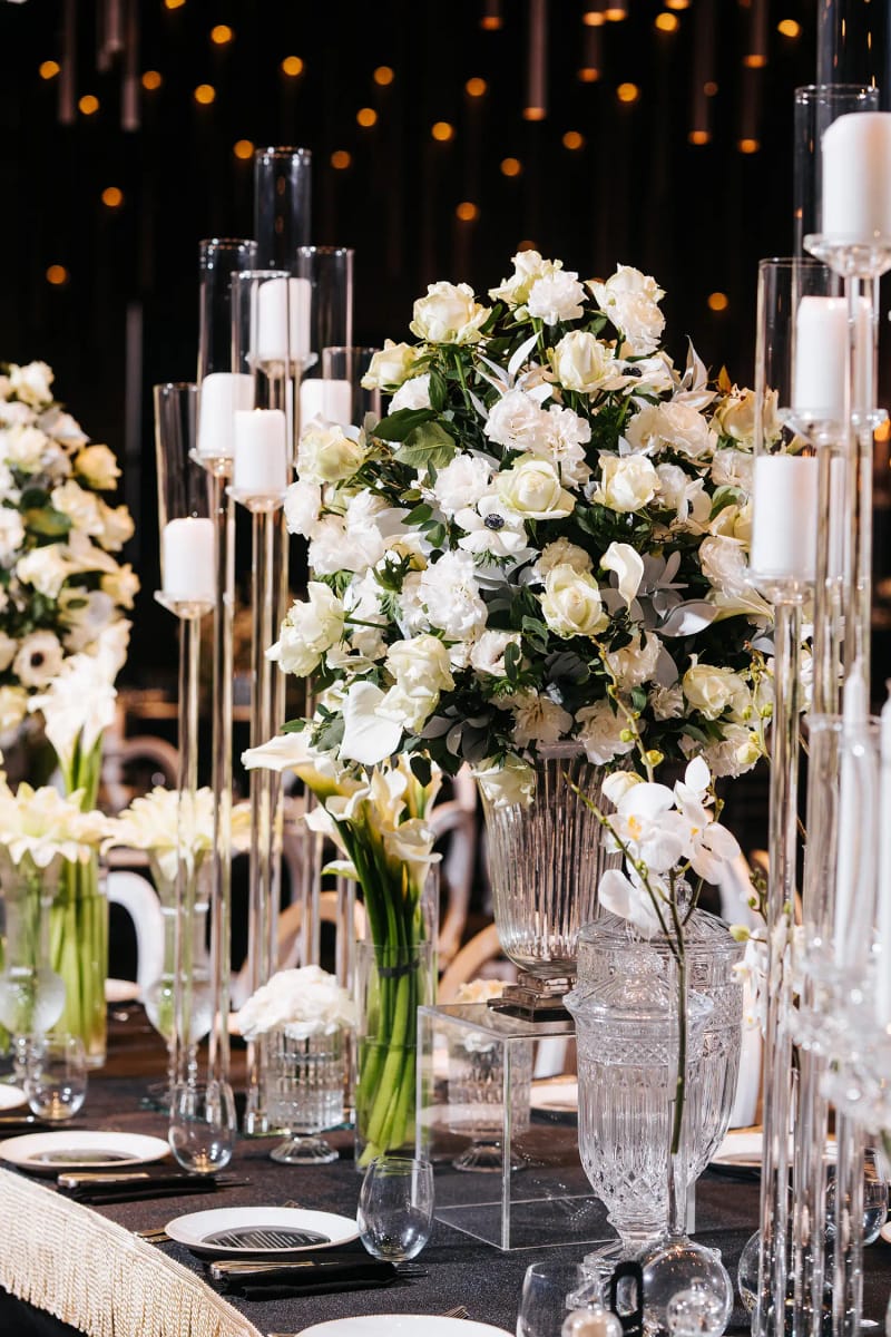 Large white rose bouquet and tall glass candle holders on a black table, with white chairs beside it