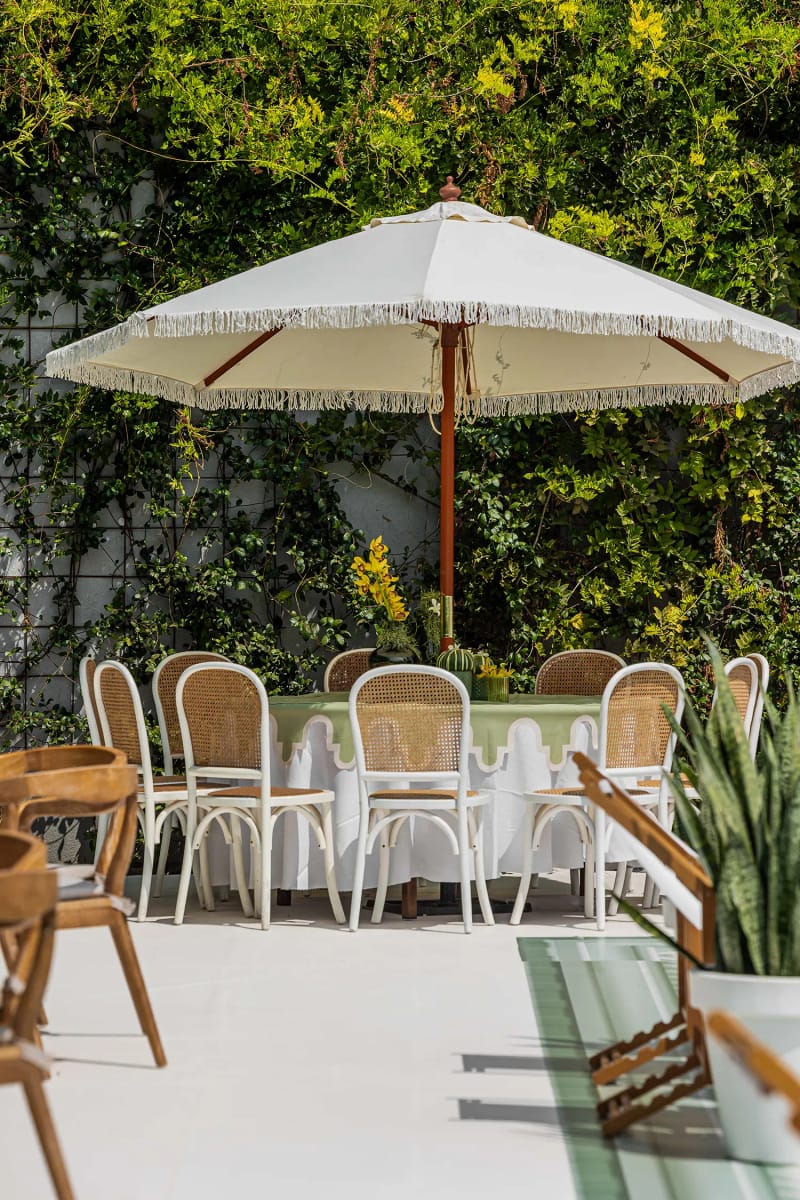 Grand parasol à franges au-dessus d'une table ronde et de chaises blanches, près d'un mur de lierre sur une terrasse claire