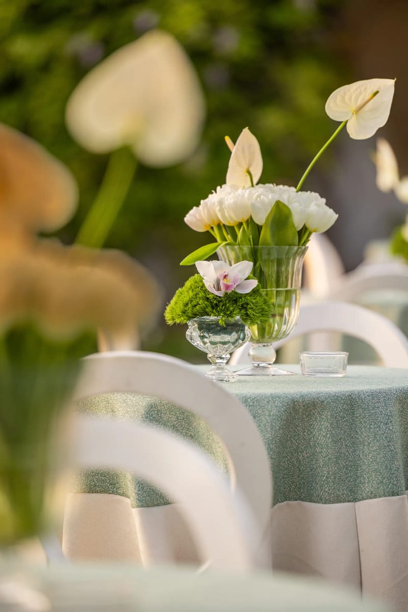 Anthurium blanc, tulipes et orchidée dans un vase en verre sur une nappe vert pâle, avec des chaises blanches floues derrière