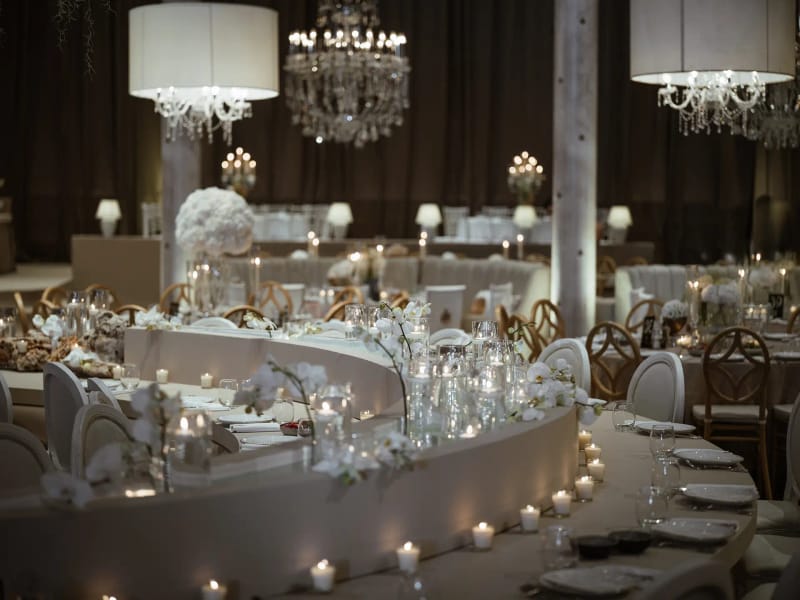 Table de banquet blanche courbe avec bougies sous des abat-jour cylindriques et des lustres suspendus, rideaux bruns au fond