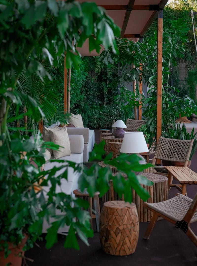 Lounge corner with a white sofa, woven chairs, a wooden stool and a lamp among plants and wooden posts