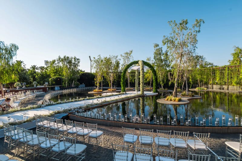 Green arch at the end of a white aisle crossing the pond under a blue sky, with rows of chairs at the side
