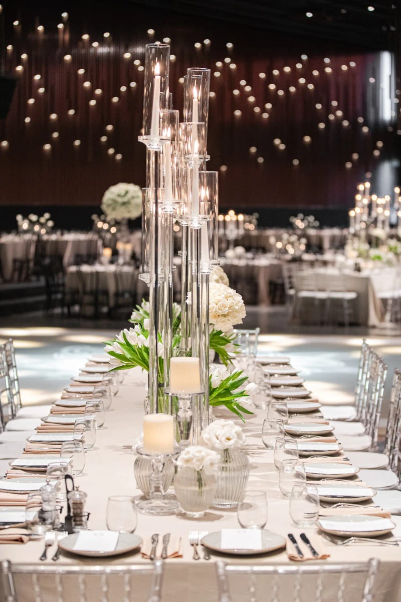 Long table with tall glass candle holders and white roses beneath a ceiling of hanging points of light