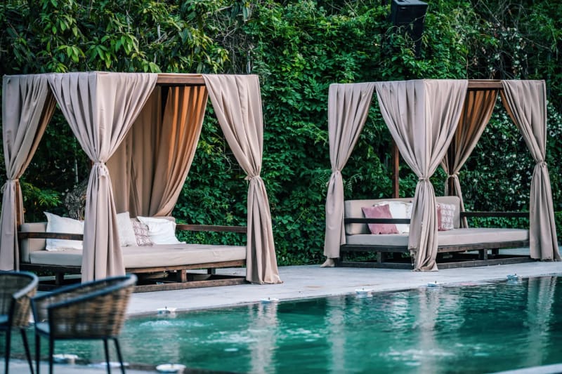 Two canopy daybeds with beige curtains against an ivy wall, with wicker chairs and a pool edge in the foreground