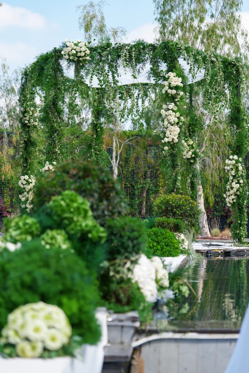 Greenery-draped canopy with white flowers beside a dark pond, with white and green blooms in the foreground