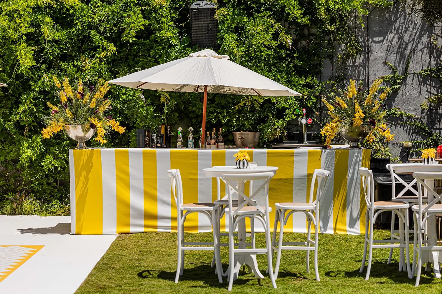 Yellow and white striped bar counter with a parasol, high tables and white chairs on the lawn among greenery