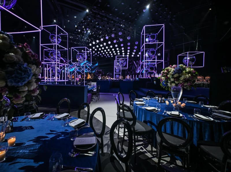 Round tables with blue cloths and black round-back chairs, neon frames, spotlights and a flower arrangement in a tall vase