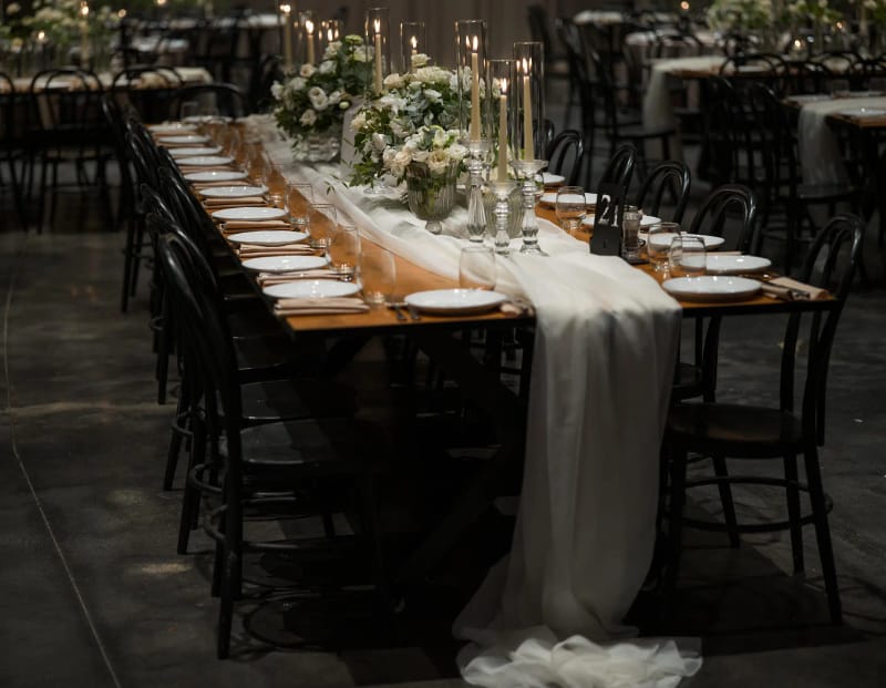 Long table with plates set, a white fabric runner draping to the floor, candles and black chairs