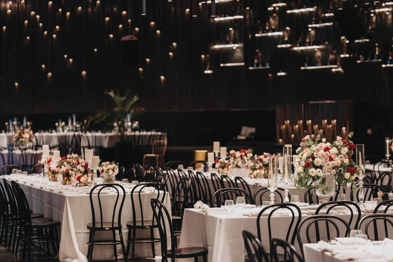 Salle aux rangées de tables, chaises noires et centres de roses rouges, petites lumières suspendues au plafond sombre