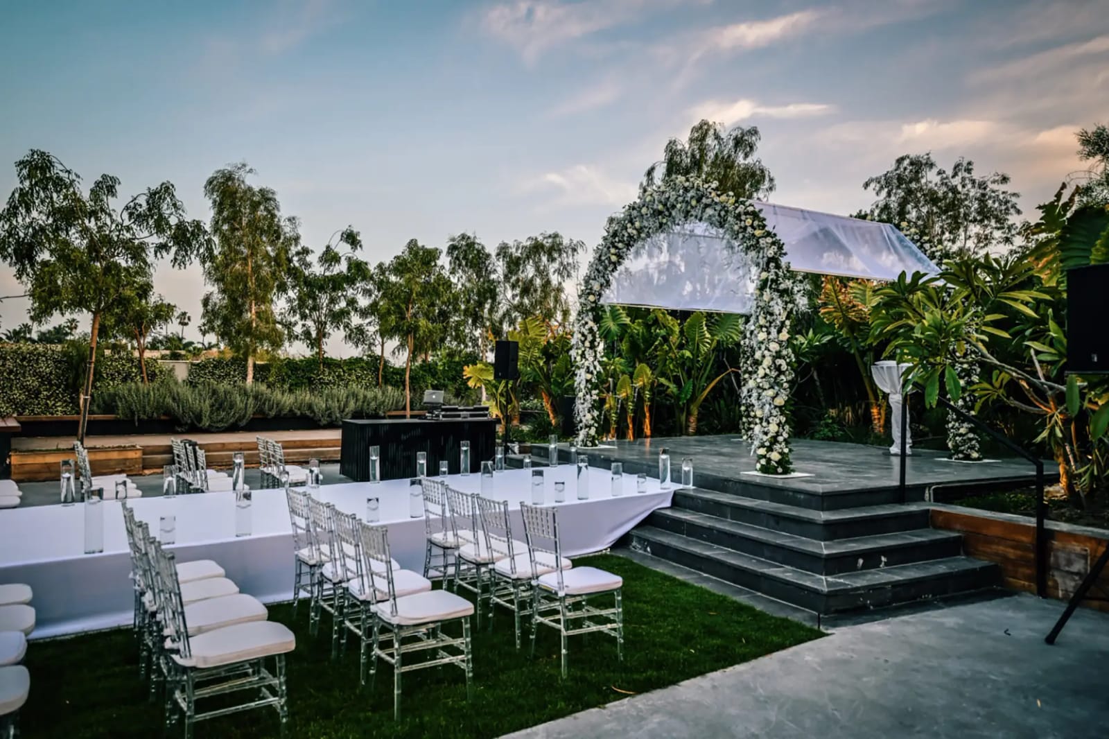 Floral arch with a white canopy on a dark stage with steps, beside linen-covered tables and clear chairs