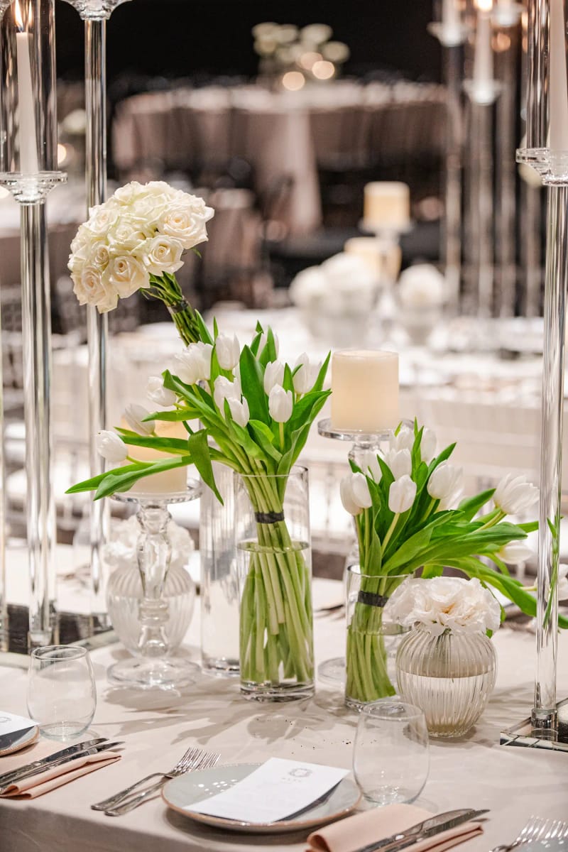 Close view of a set table with a white rose in a tall glass holder, calla leaves in glass vases and a menu card on a plate