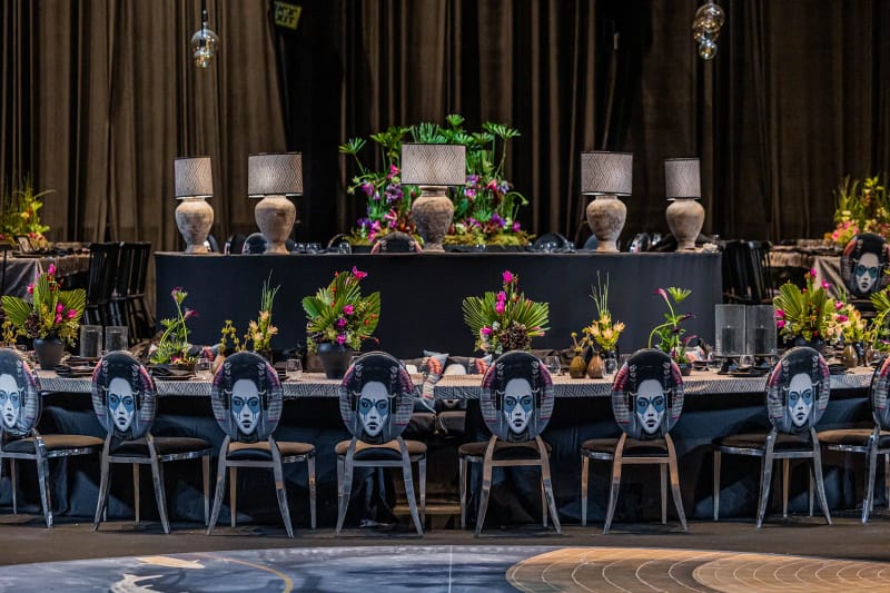 Long table with chairs whose backs carry portraits, stone-based lamps and plant arrangements before curtains