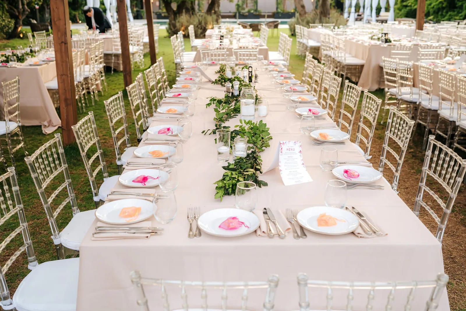 Longue table de banquet avec chemin de verdure, assiettes ornées de pétales roses et chaises transparentes des deux côtés