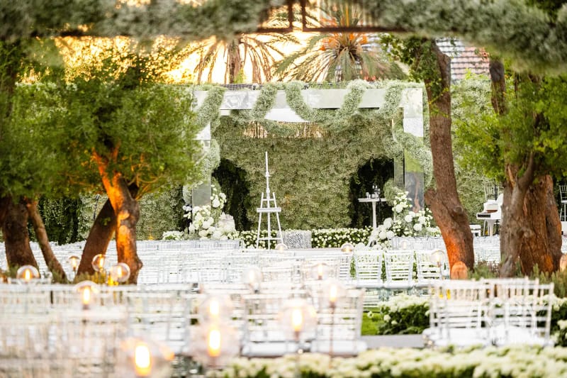 Flower and greenery backdrop with an easel between lit olive trees, with candles and white chairs in front