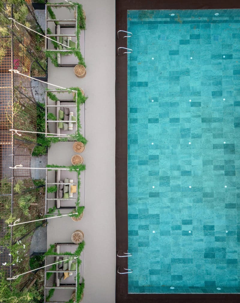 Overhead view of a long turquoise-tiled pool beside a row of seating nooks dressed with greenery