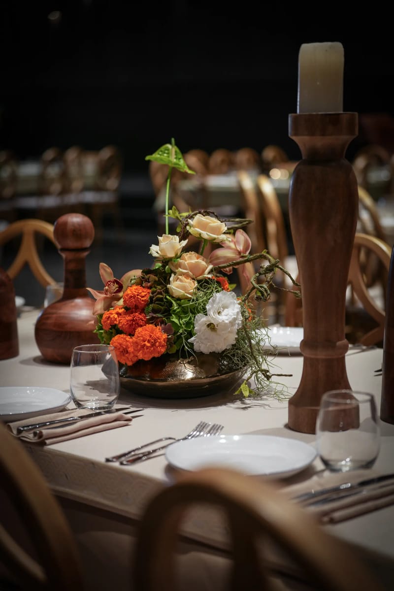 Arrangement of orange and white flowers in a shallow bowl beside a carved wooden candlestick and wooden bottles
