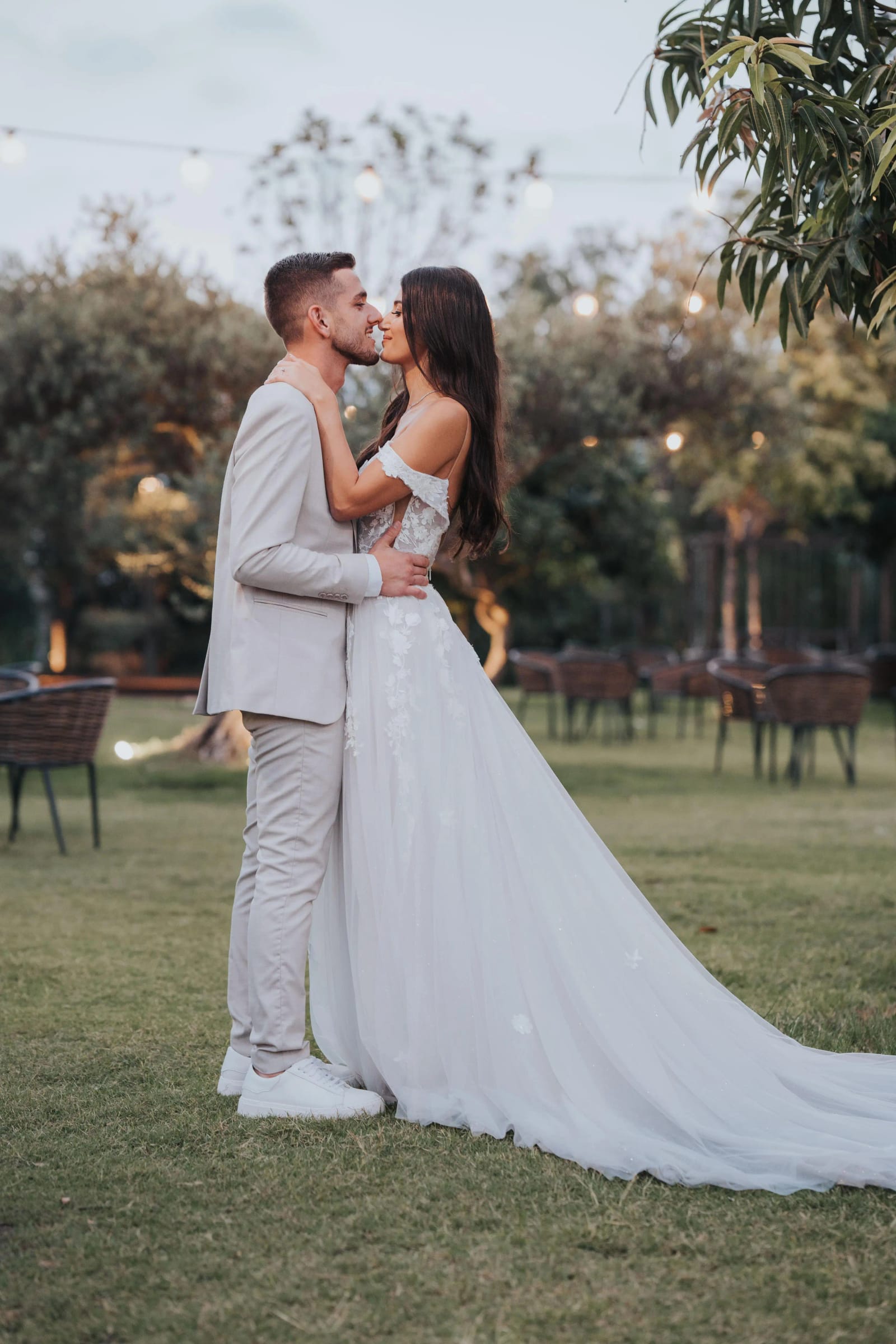A couple kissing on a lawn, the bride in a gown with a long train, with garden trees and string lights behind