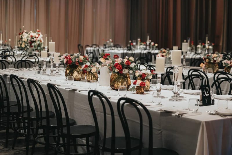 Longue table nappée de blanc avec chaises bistrot noires, vases dorés de roses rouges et roses et bougies pilier