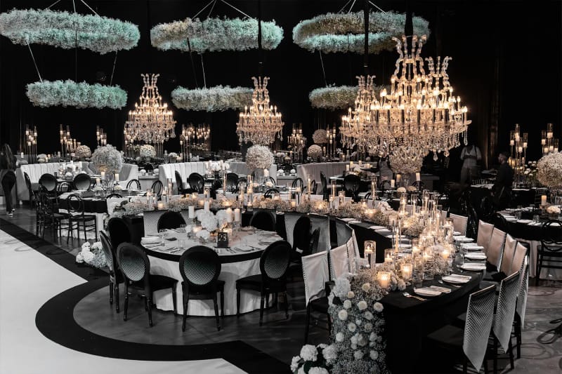 Round and curved banquet tables under crystal chandeliers and hanging floral rings in a dark hall at Hangar