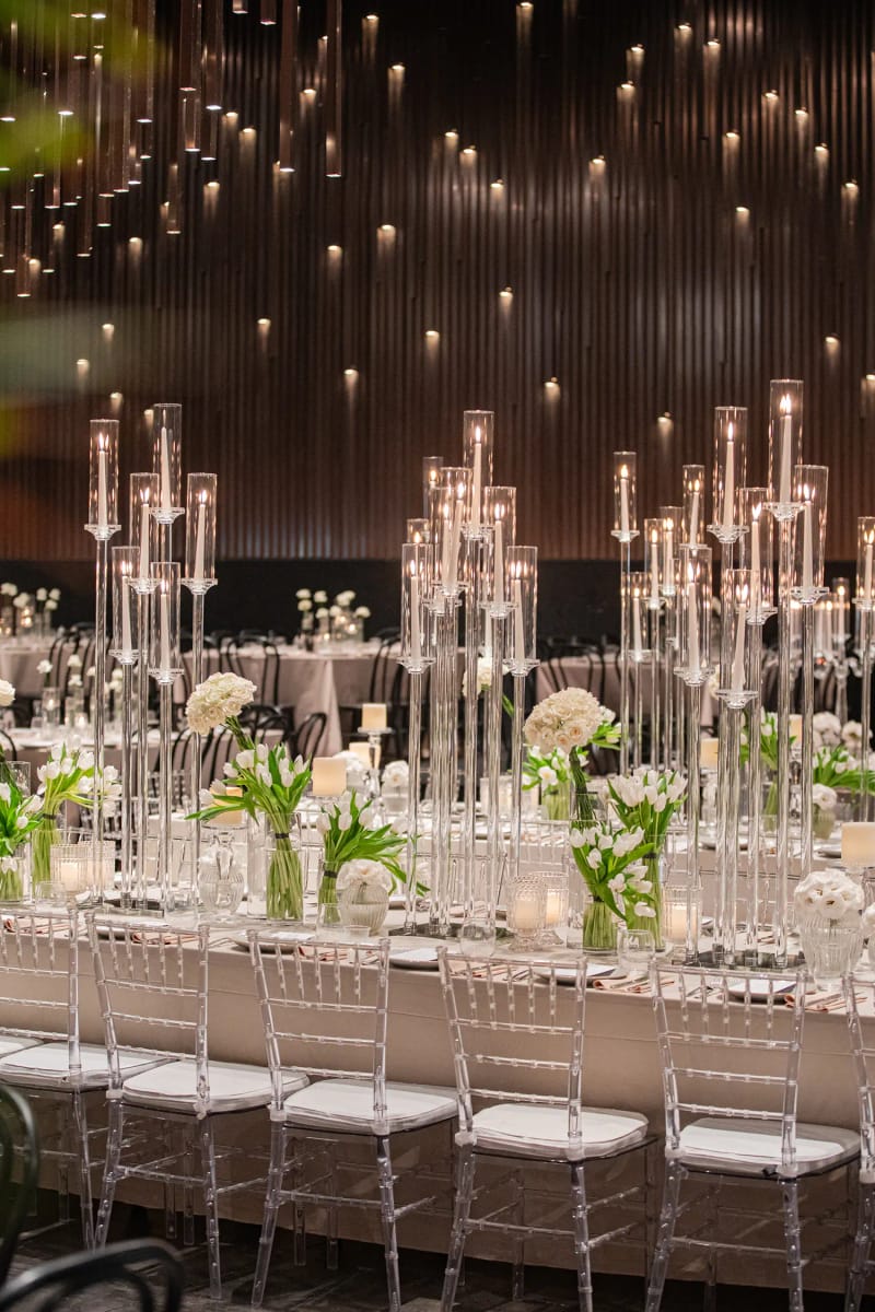 Tall glass candelabras on a long table with white flowers and clear chairs, under a ceiling of small lights