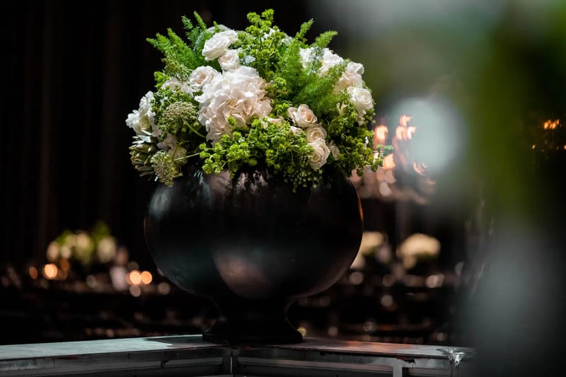 Vase rond noir avec composition de fleurs blanches et vertes et de fougère, devant une salle sombre floue