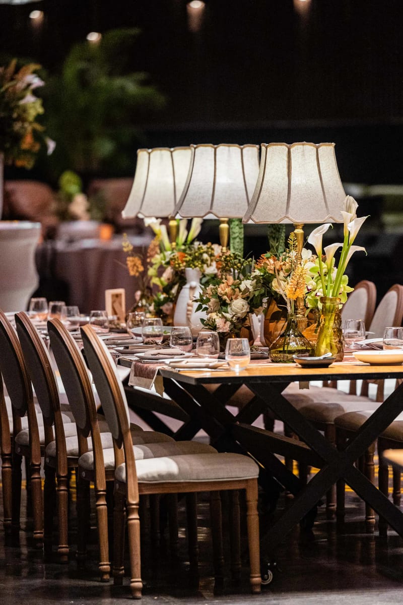 Longue table en bois à pieds croisés avec lampes et fleurs, chaises de chaque côté dans une salle sombre