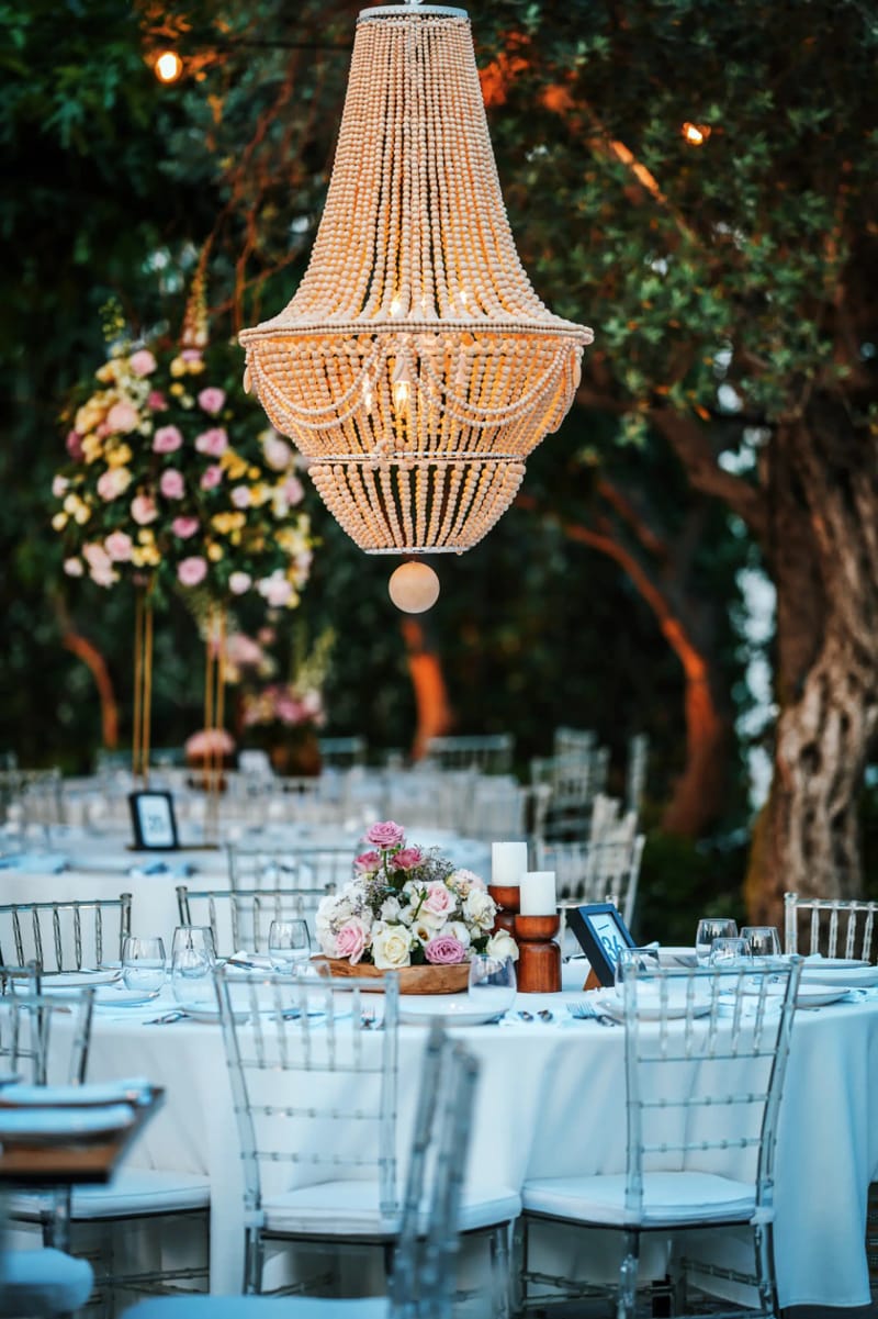 Beaded chandelier hanging from a tree above round tables set with flowers and clear chairs