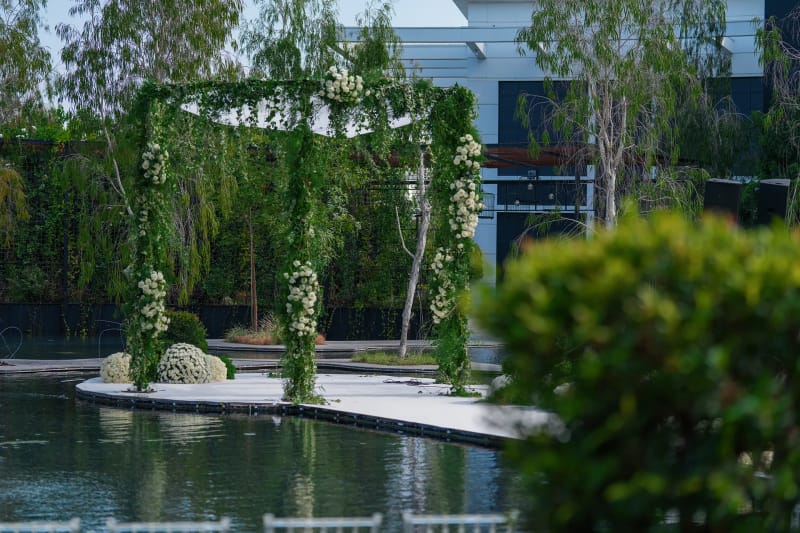 Greenery-wrapped canopy over a white platform in a dark pond, with birch trees and a modern building behind