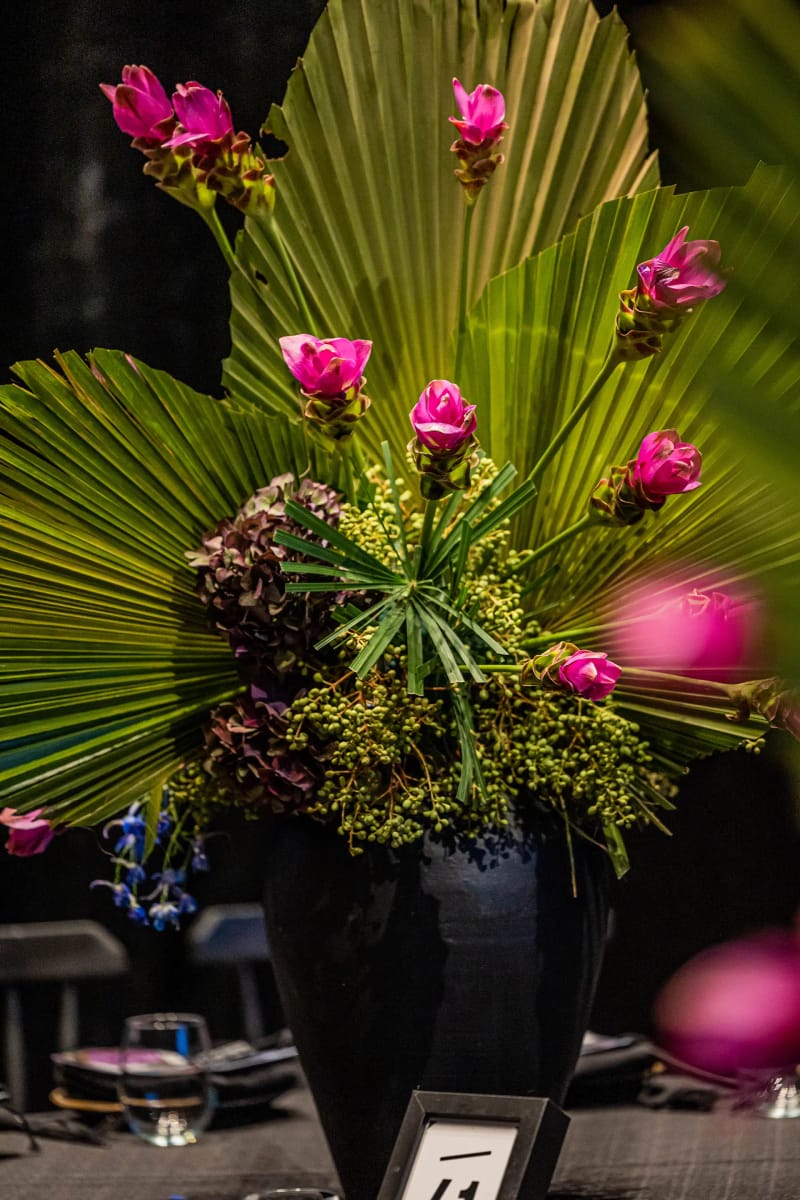 Tall arrangement of fan palm leaves and pink flowers in a black vase with a table number card at its base