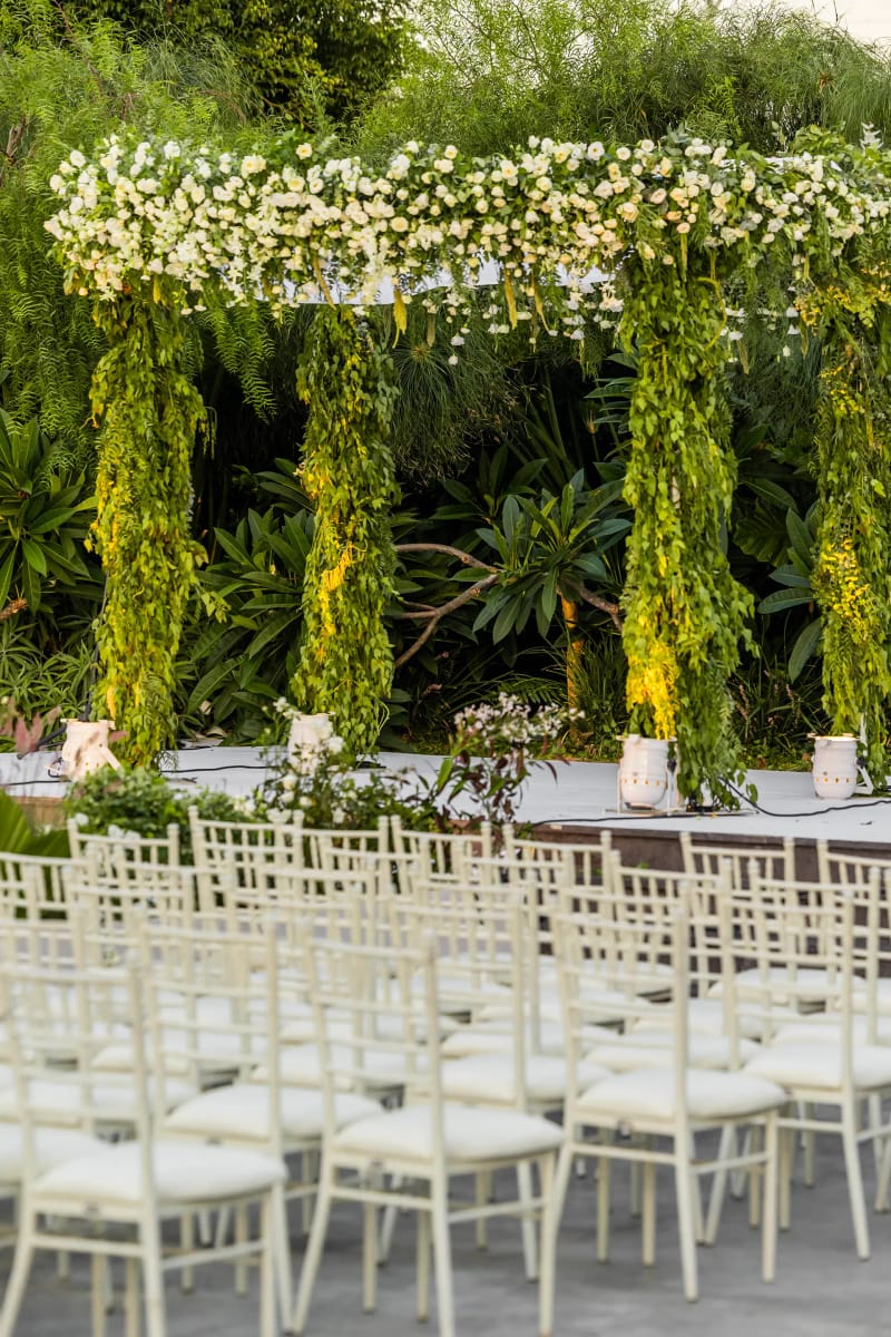 Rows of white chairs facing a rectangular flower-topped canopy on greenery-wrapped posts in a green garden