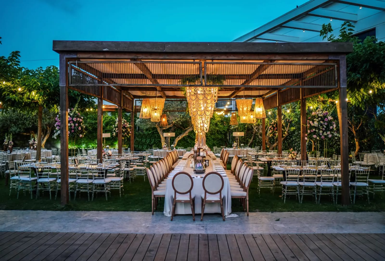 Wooden pergola with chandeliers over a long table at dusk, with round tables on the lawn and a wooden deck in front