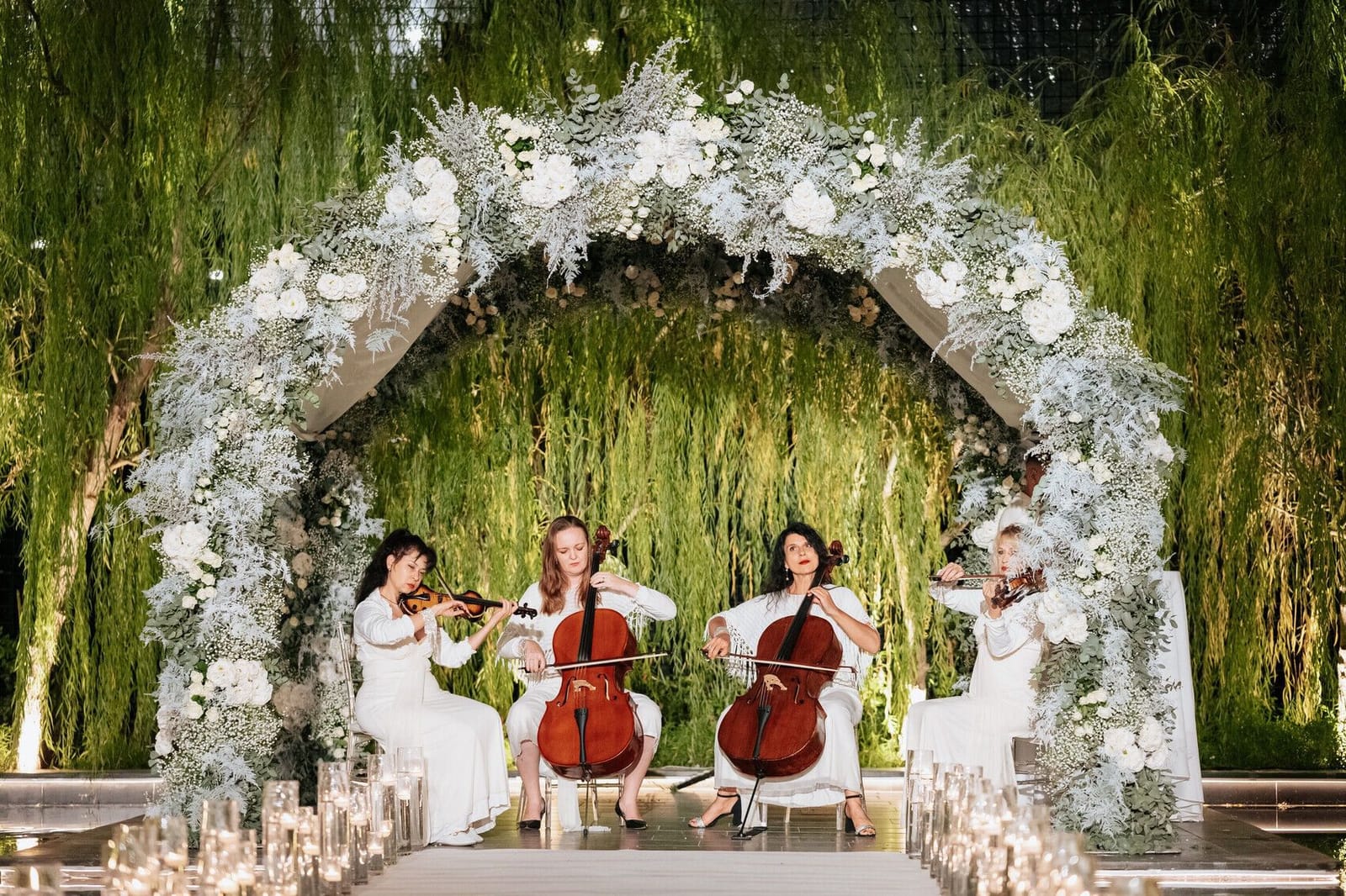 Un quatuor à cordes en blanc jouant violons et violoncelles sous une arche fleurie, devant des saules et des bougies