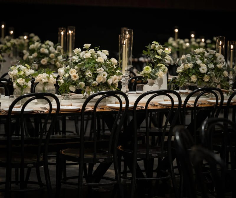 White flower arrangements with foliage beside tall candles in glass cylinders on a table in low light