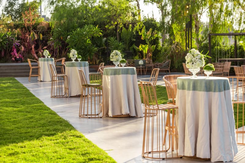Cocktail tables with pale cloths and gold wire stools along a stone path and lawn in a garden with string lights