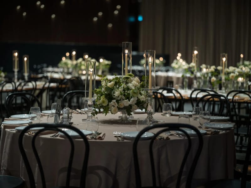 Round table with a taupe cloth, a green-and-white floral centerpiece and tall candles, surrounded by black chairs