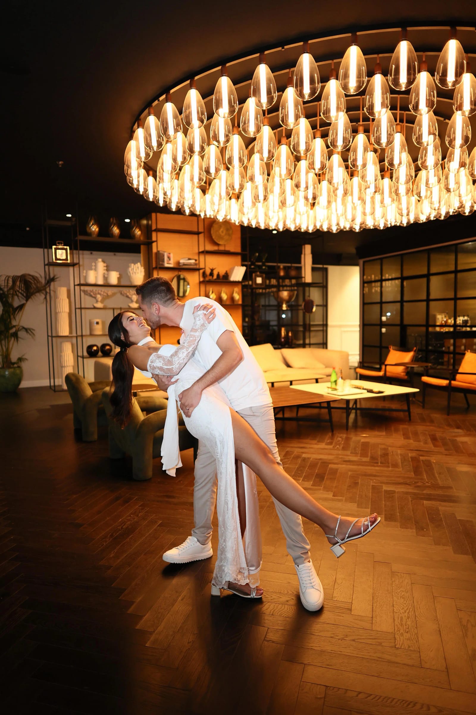 A couple sharing a dipped kiss in a warm lounge beneath a huge oval bulb chandelier, with wooden shelves behind