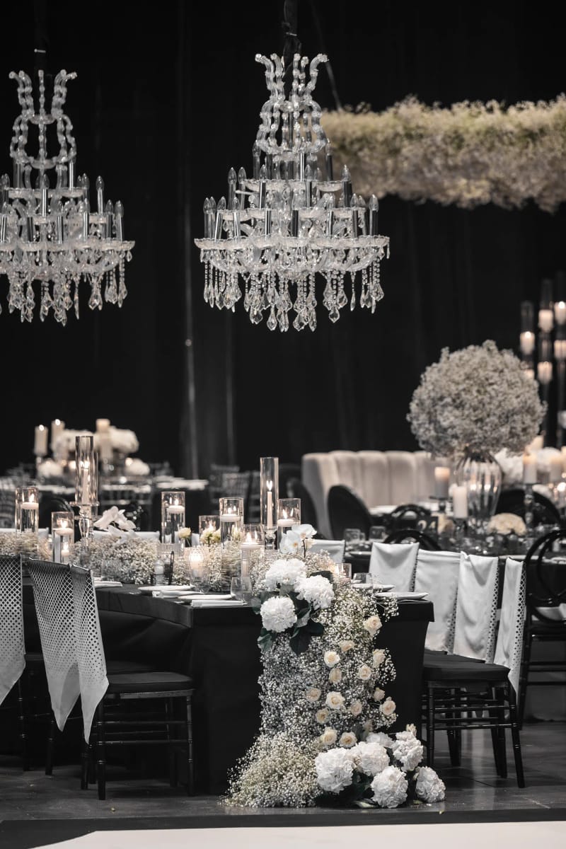Lustres en cristal au-dessus d'une longue table ornée d'un chemin de fleurs blanches en cascade, tables rondes au fond