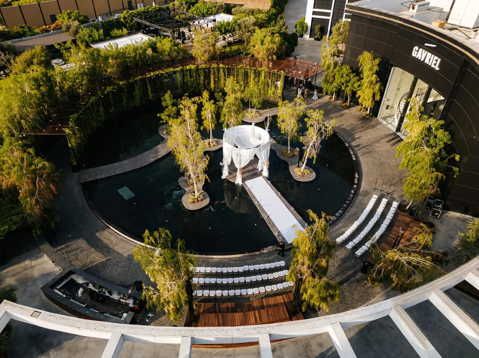 Vue aérienne d'un bassin rond sombre aux îlots plantés d'arbres, dais blanc au bout d'une allée blanche, chaises en arc