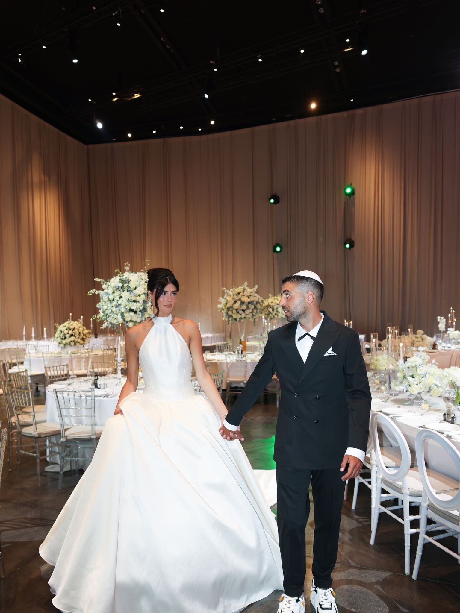 A couple standing among tables with white floral centerpieces, in a hall with wood-paneled walls and spotlights