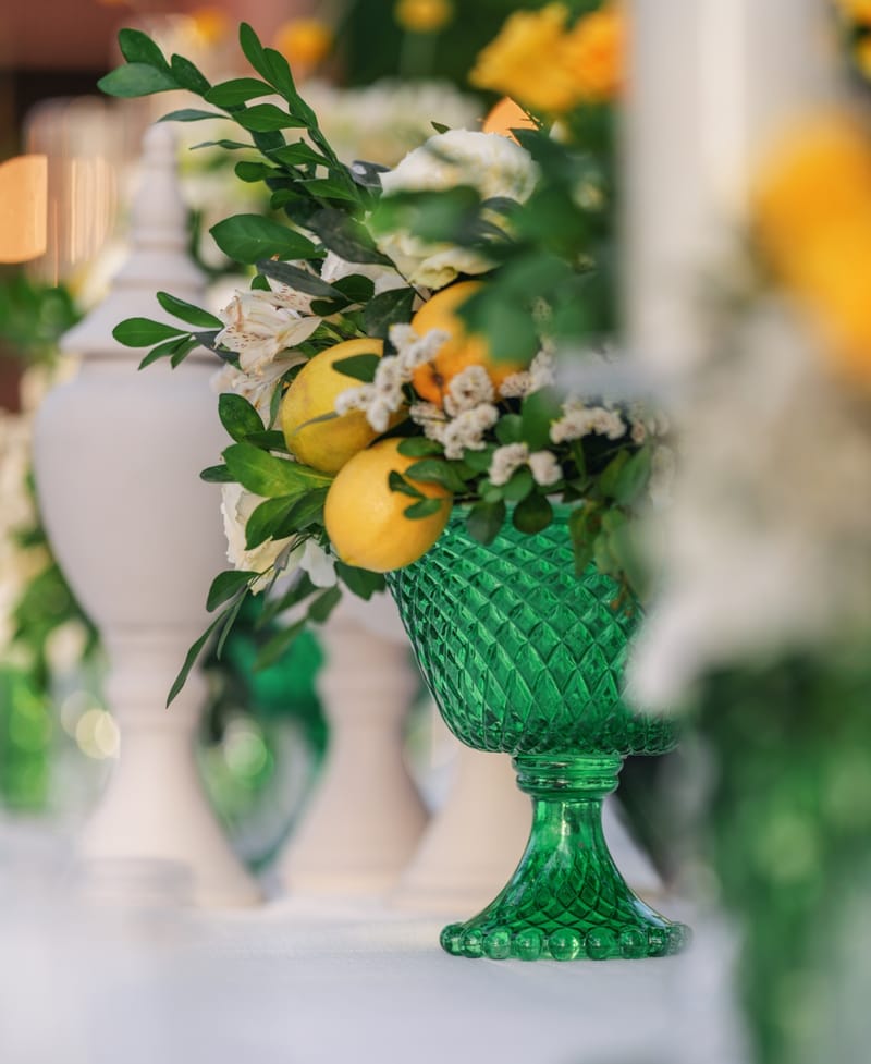 Close-up of a green cut-glass urn with flowers and lemons in front of a white ceramic urn