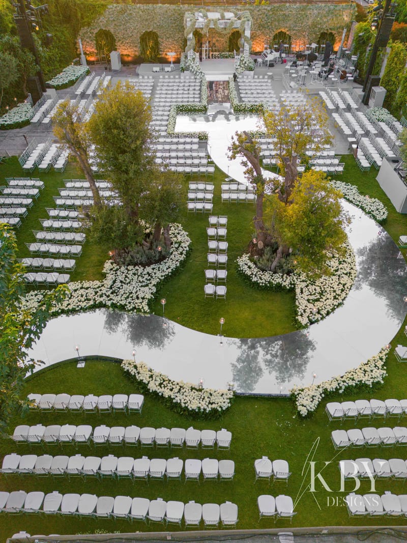 Aerial view of rows of white chairs on a lawn around a curving mirrored aisle with trees and white flowers