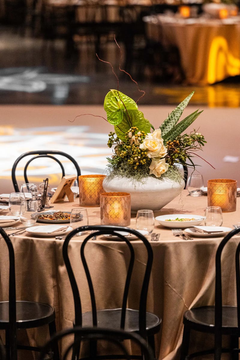 Table ronde à nappe champagne, chaises bistrot noires, bougeoirs ambrés et roses crème dans une coupe blanche
