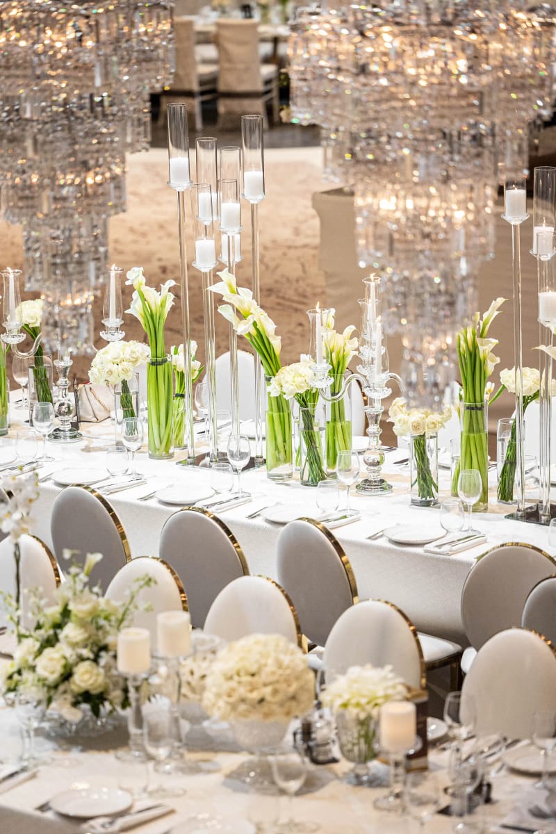 Lustres de cristal étagés au-dessus d'une longue table blanche avec callas dans des vases en verre et chaises blanches