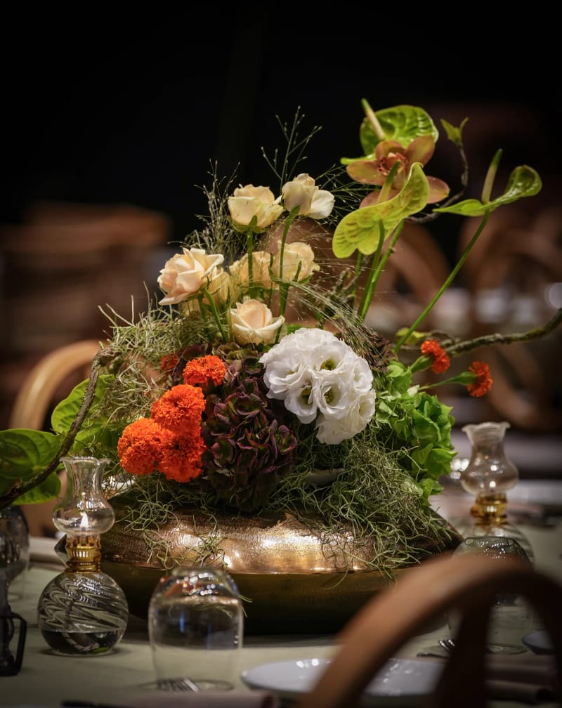 Close-up of orange, white and green flowers with moss in a brass bowl, flanked by small glass lanterns